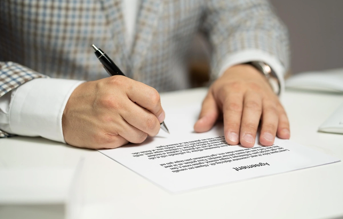 Close-up of a person in a checked blazer signing a printed agreement on a white desk with a black pen