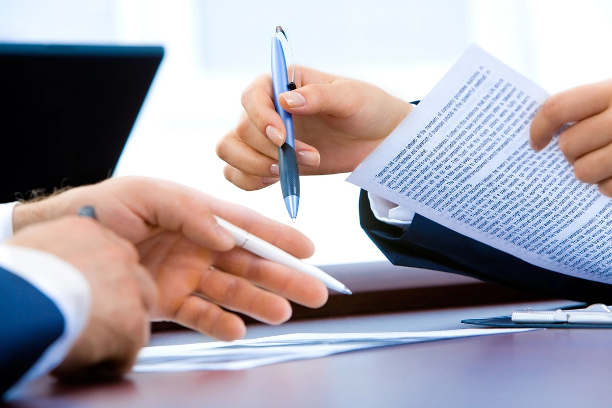 Two people at a desk going over a printed document with pens, a laptop open beside them