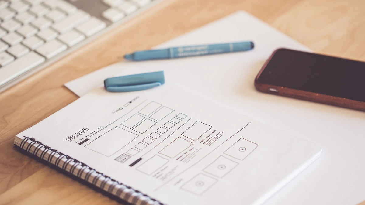 A spiral notebook with hand-drawn website wireframe sketches on a wooden desk next to a keyboard, a blue marker, and a phone
