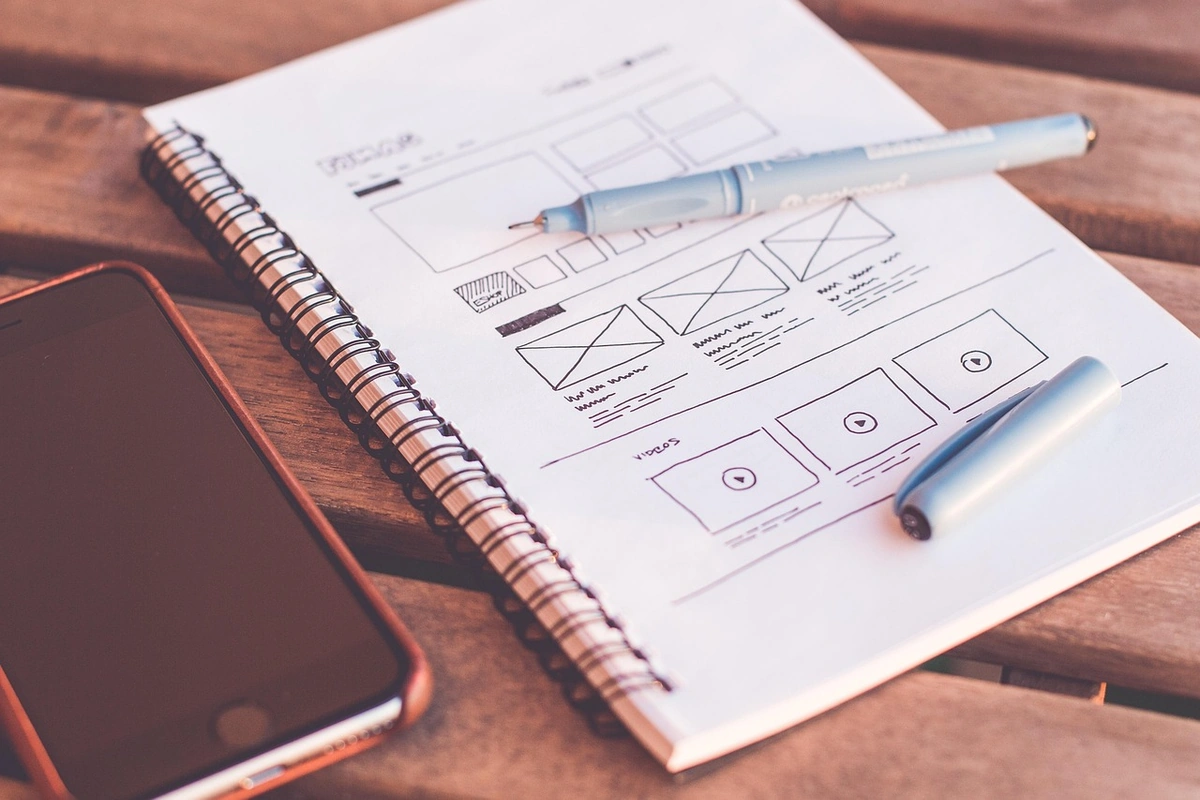 A hand-drawn website wireframe sketched in a spiral notebook on a wooden table, with two pens and a smartphone beside it