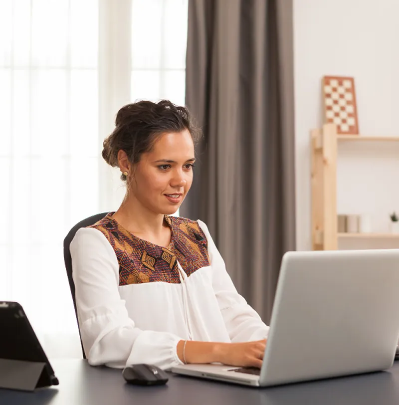 Woman in a white blouse with patterned details working on a laptop at a desk in a bright room.